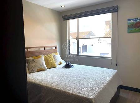 Bedroom with double bed, wood headboard, yellow decorative pillows, large window with gray frame overlooking rooftops, recessed ceiling light.