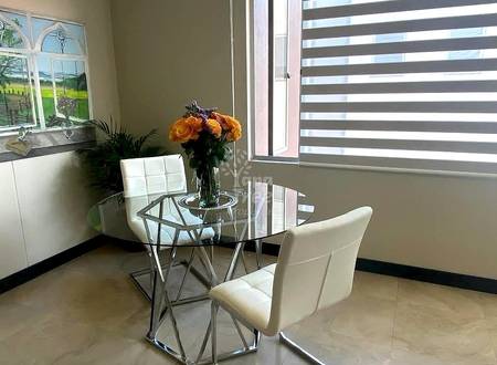Bright living room with round glass table, cream chairs, orange flowers in vase, striped window blinds, and tile flooring.