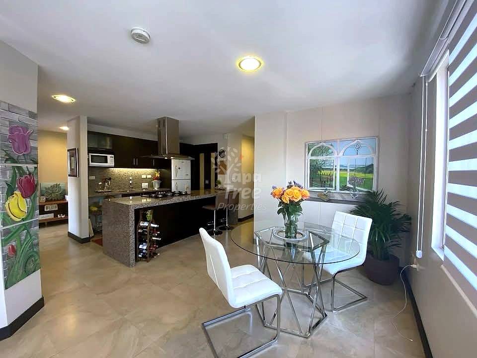 Open-concept departamento with black kitchen island, dining area with round glass table and white chairs, recessed ceiling lights, and striped window blinds.