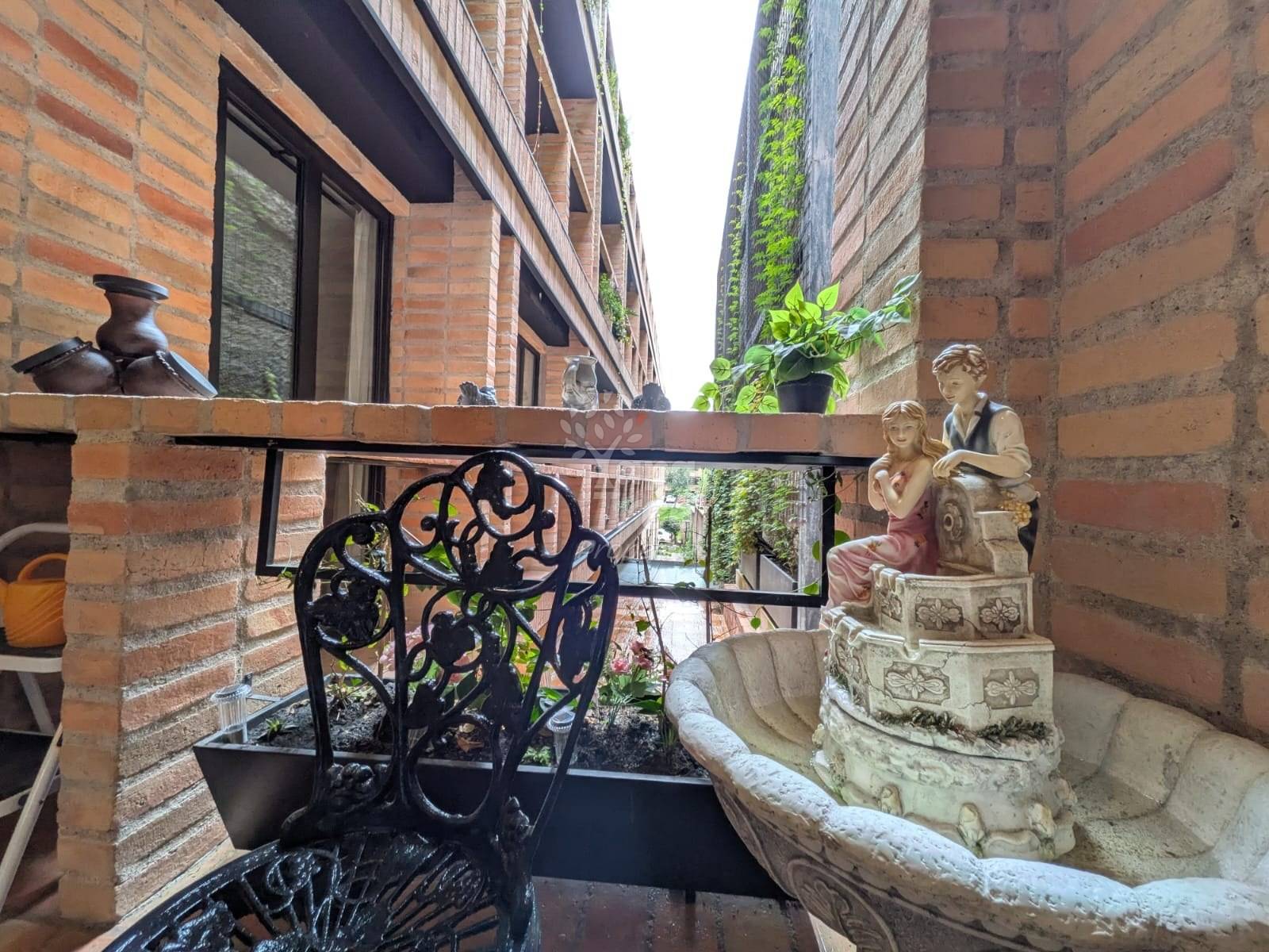 Narrow balcony with exposed red brick walls, black wrought iron railing, decorative statue, and potted plants overlooking adjacent building