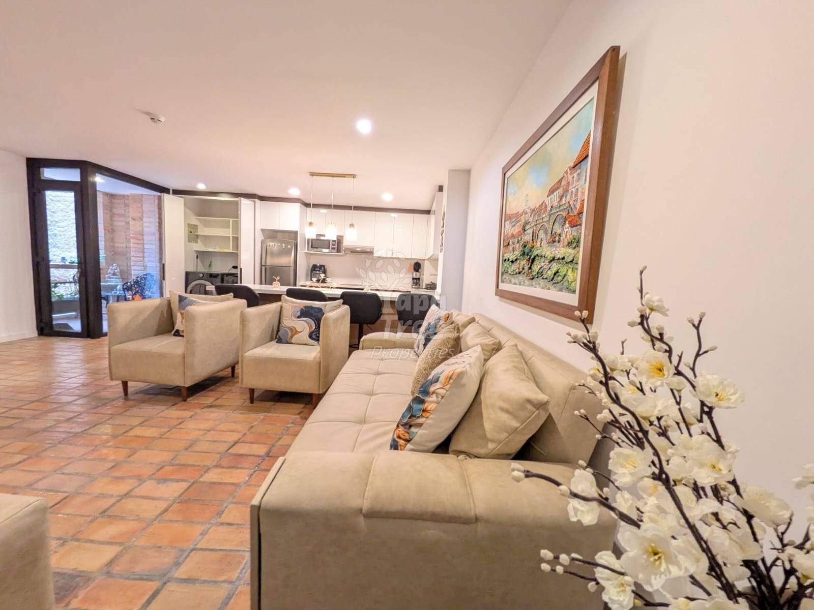 Open-plan living room with cream sectional sofa, terra cotta tile flooring, white walls, recessed lighting, and kitchen visible in background with dark wooden doors.