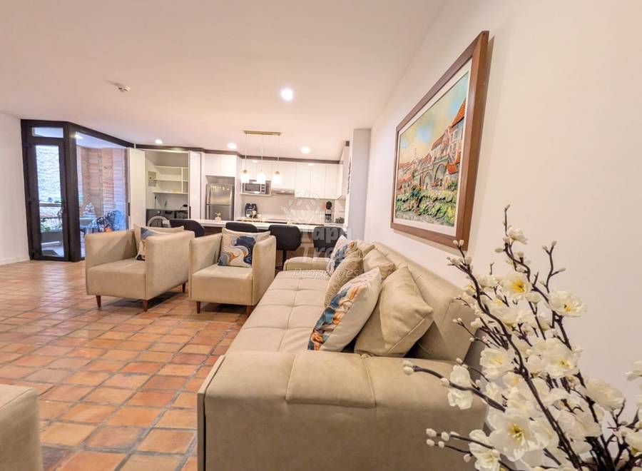 Open-plan living room with cream sectional sofa, terra cotta tile flooring, white walls, recessed lighting, and kitchen visible in background with dark wooden doors.